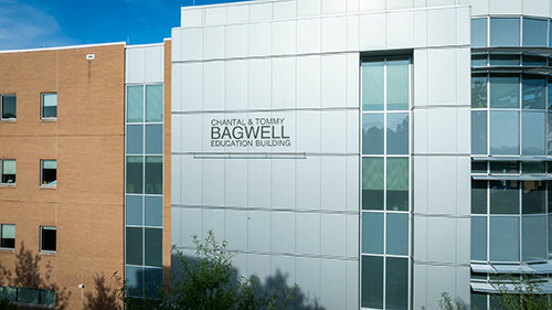 A modern educational building with a mix of brick and metal facade. The name 'Chantal & Tommy Bagwell Education Building' is prominently displayed on the metal section of the building. The sky is clear and blue in the background.
