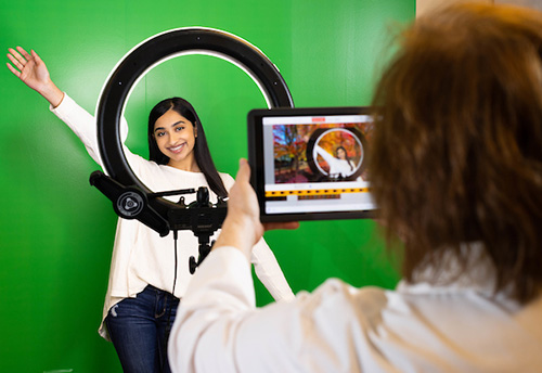 A student captures a photo with her camera, positioned in front of a vibrant green screen backdrop.