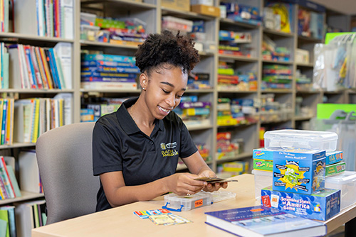 A student sitting at a library table, focused on her work, with bookshelves filled with literature around her.