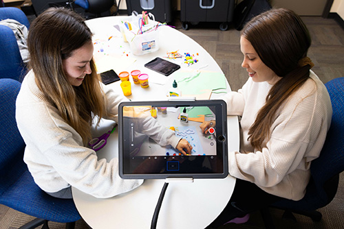 Two students engaged in conversation while sitting at a table, using an iPad for interaction or entertainment