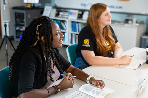 Two students engaged in discussion at a table, surrounded by books and pens, illustrating a collaborative study environment.