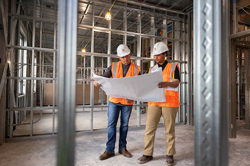 Two men wearing hard hats stand in a construction area, observing the ongoing work and discussing project details.