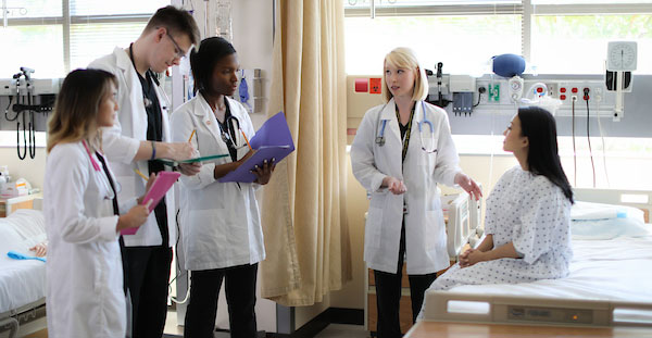 In a hospital room, a patient is conversing with a doctor as students take notes and learning from the KSU Wellstar College of Health and Human Services.