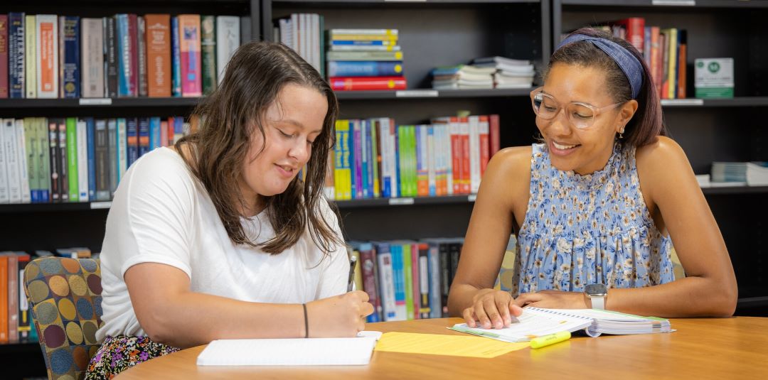 A KSU student and instructor with paper and a notepad, sitting at a desk, surrounded by books and study materials.