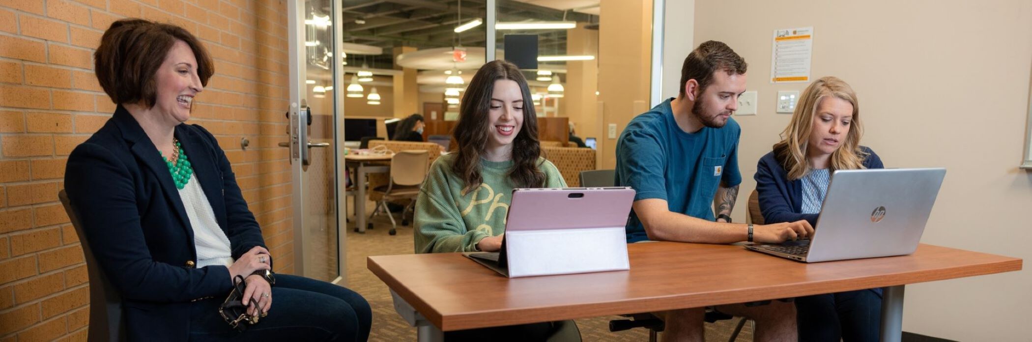 A small group setting with a professional (advisor) and students collaborating around a table, using laptops and tablets.