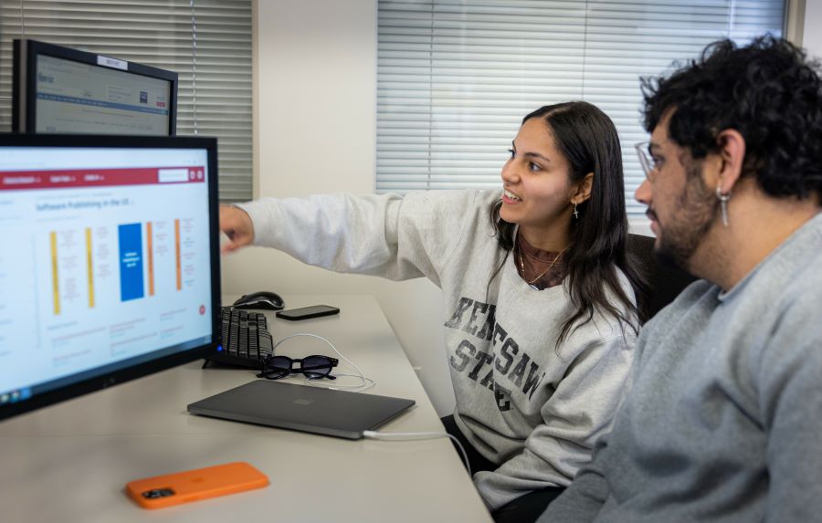 Two students collaborating at a desk with a computer screen displaying data.