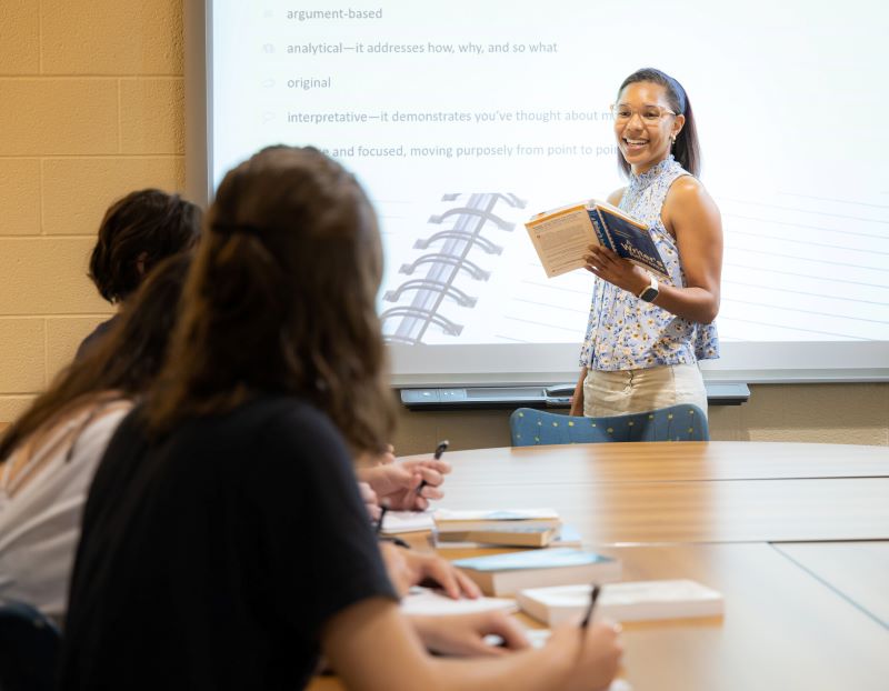 A teacher stands at the front of a classroom, smiling and holding a book, as students seated at a table listen and take notes. A presentation is projected in the background.