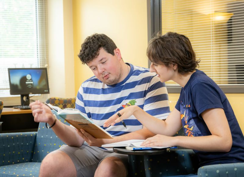 Two students collaborate on a reading assignment, discussing a book while seated in a lounge area.