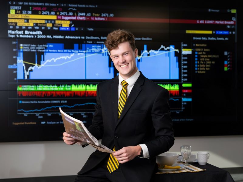 A young man in a suit sits at a desk, holding a newspaper, with a screen displaying market data in the background.