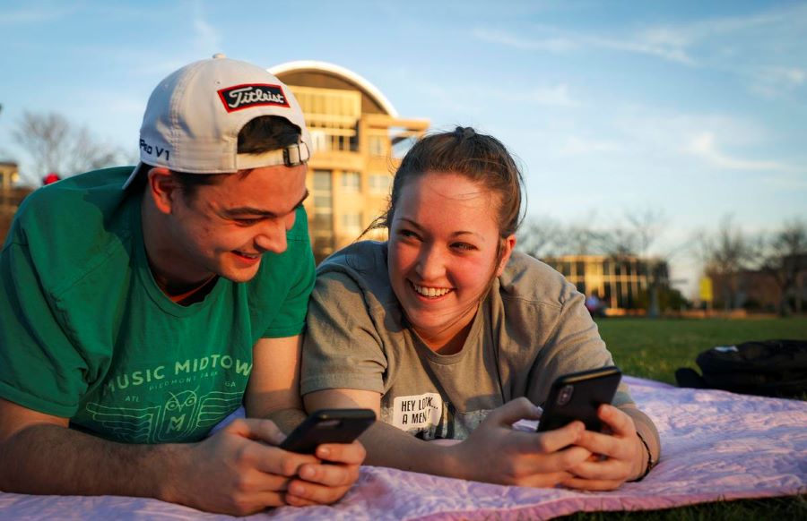Two KSU students lie on the grass, focused on their phones, surrounded by a serene outdoor environment.