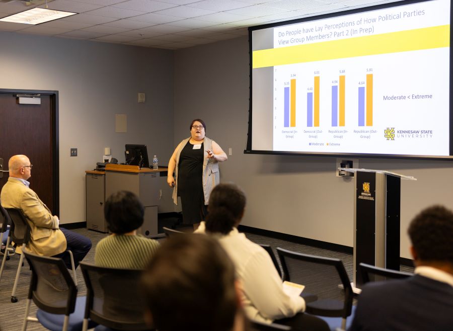 A woman presents to an audience, with a chart displayed on the screen behind her, illustrating key data points.
