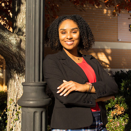 A woman in a suit and red shirt leans against a pole, representing professionalism at Coles College of Business.