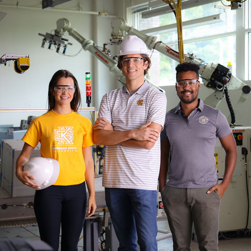 Three KSU students in yellow shirts and hard hats pose in front of a machine, symbolizing teamwork in engineering and math