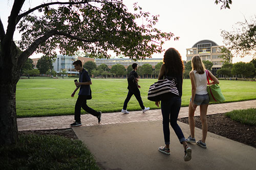 Three Kennesaw State students walk on a sidewalk beside a grassy area, with Kennesaw Hall visible in the background.