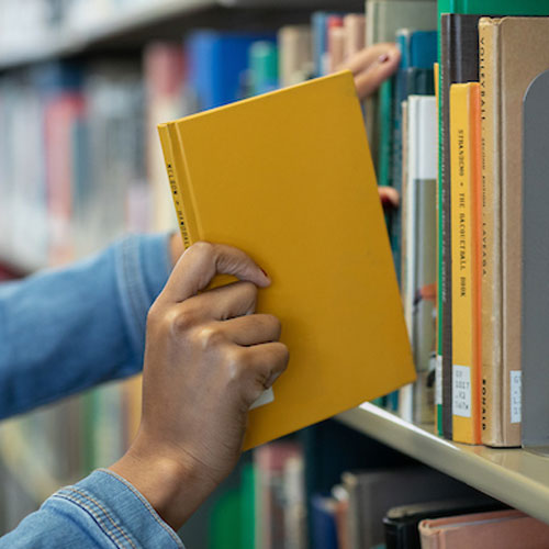 A library professional at the College of Humanities holds a book on a shelf, surrounded by numerous other books.