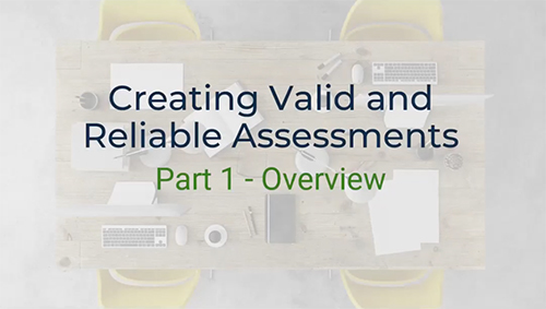 A top-down view of a conference table with a laptop, keyboard, mouse, pens, and papers scattered across it. The text "Creating Valid and Reliable Assessments&" and "Part 1 - Overview" is written on the table. Two yellow chairs are visible on either side of the table.