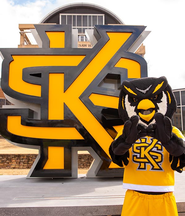 scrappy taking a picture on campus in front of the ksu sign.