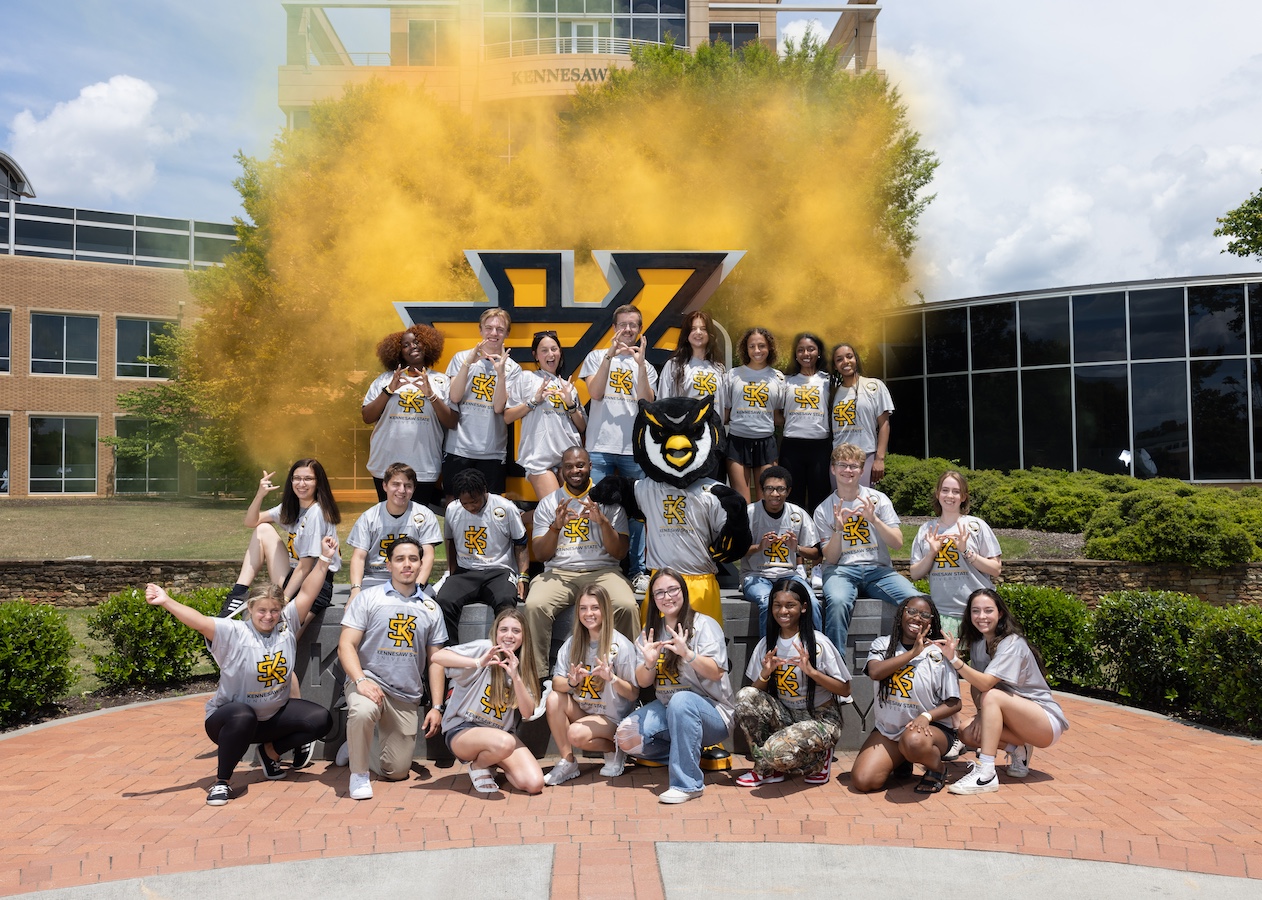 Large group of first year students excitedly taking a picture with scrappy in front of the ksu sign on campus.