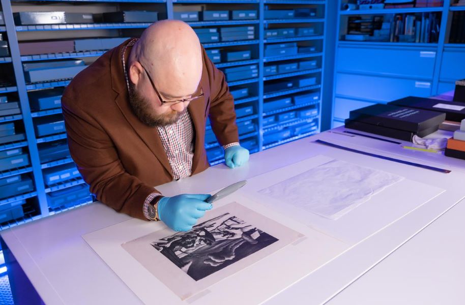 Person examining a black-and-white print with a magnifying glass in a storage room