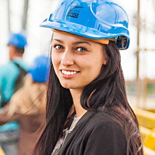 A KSU student wearing a blue construction hard hat and a black jacket smiles at the camera while standing on a construction site.