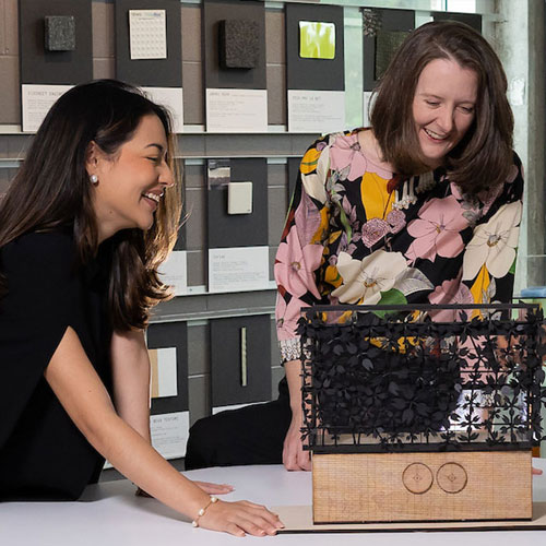 Two women smiling and examining an architectural model made of wood and intricate black cut-out material, set on a table in front of a wall covered with sample materials and swatches.