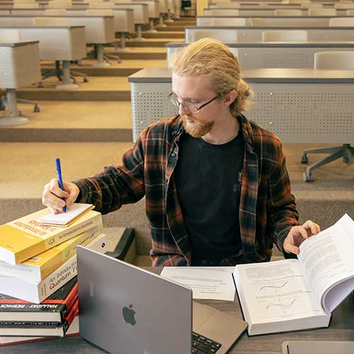 csm student sitting at a desk with their laptop and a stack of books, taking notes.