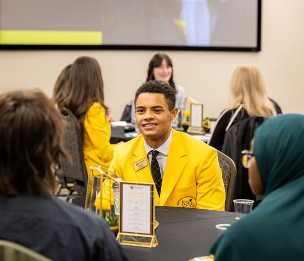 People seated at a table during an event, with some wearing yellow jackets and a screen in the background.