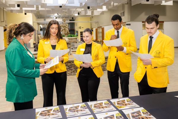 Five people indoors, four in yellow KSU blazers and one in green, looking at papers around a table