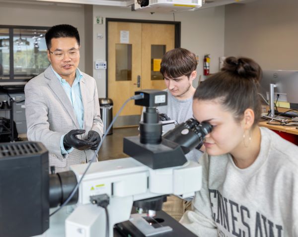Three people in a lab, two using a microscope and one standing with gloves