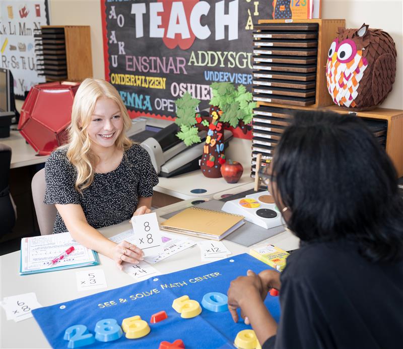 female with blonde hair smiling as she looks at another student across classroom desk