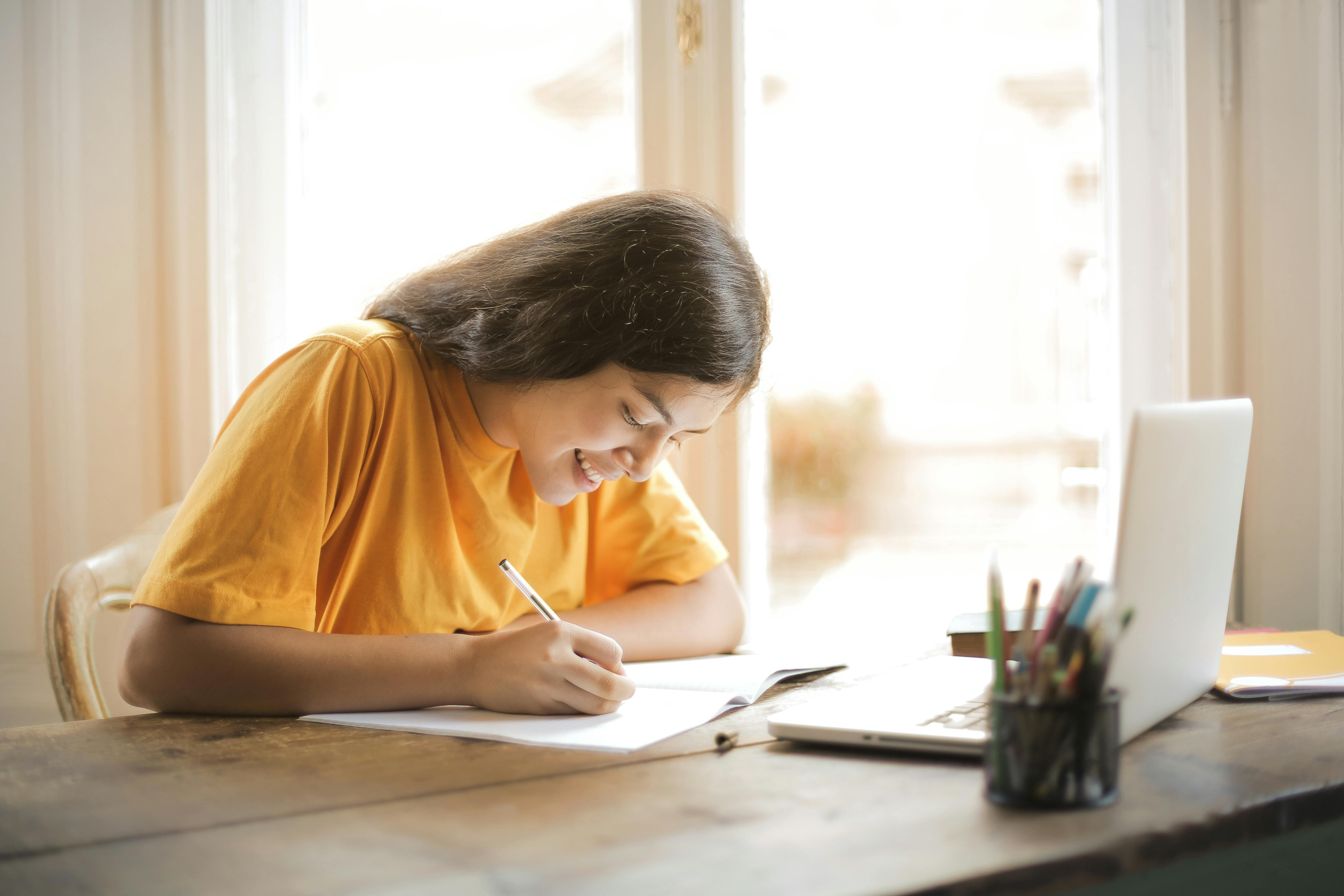 female student in yellow shirt smiling and writing on desk