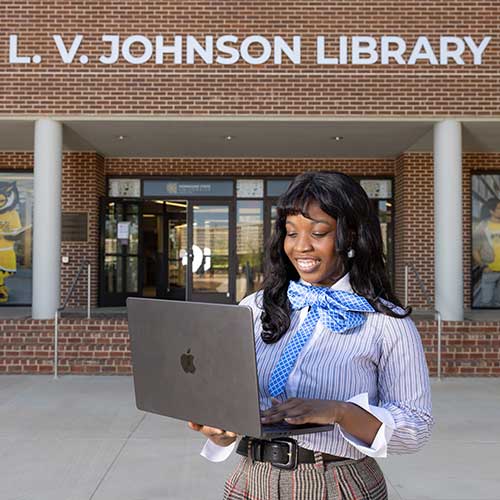 ksu student standing in front of the l.v. johnson library holding their laptop.