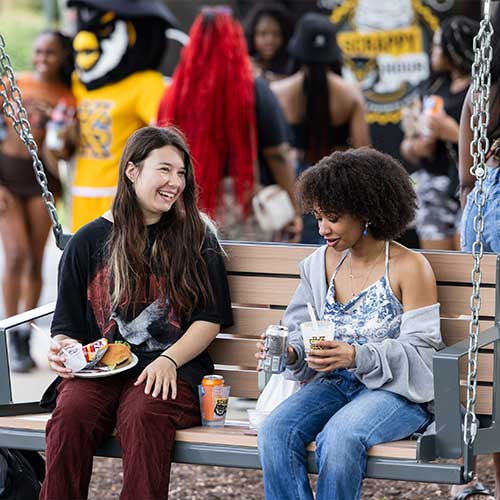 two students sitting on a wooden swing on campus during a outside event.
