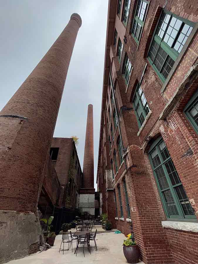 Exterior H Bldg. Condominium Courtyard (née Mill Bldg. 1), Smoke Stacks. 6/29/2022. Photograph by Nina Elsas. Courtesy of The Patch Works Art & History Center. / Exterior H Bldg. Condominium Courtyard (née Mill Bldg. 1), Smoke Stacks. 6/29/2022. Photograph by Nina Elsas. Courtesy of The Patch Works Art & History Center. (née Mill Bldg. 1), Smoke Stacks. 6/29/2022. Photograph by Nina Elsas. Courtesy of The Patch Works Art & History Center.