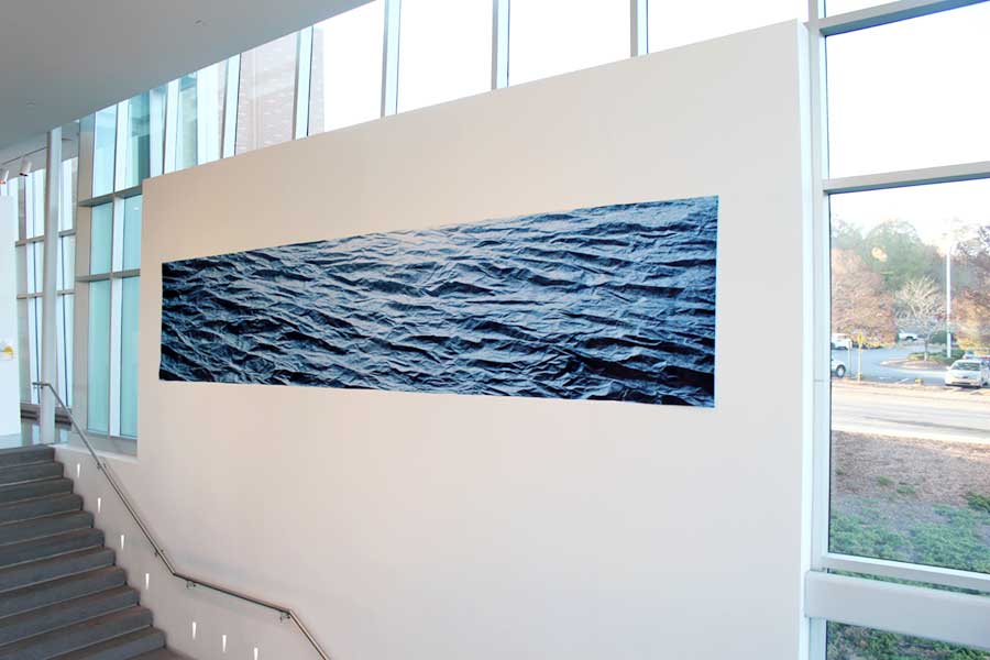 Installation view of Ben Butler's "Uncharted" which is a long rectangular piece which shows a zoomed in photo of crumpled paper, printed in blue, resembling waves of water