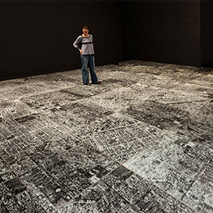 Person standing on Oscar Muñoz's "Ambulatorio (Walking Place/Outpatient Ward)" a floor piece consisted of several tiles of encased broken glass on top of a black and white photograph of Cali Columbia