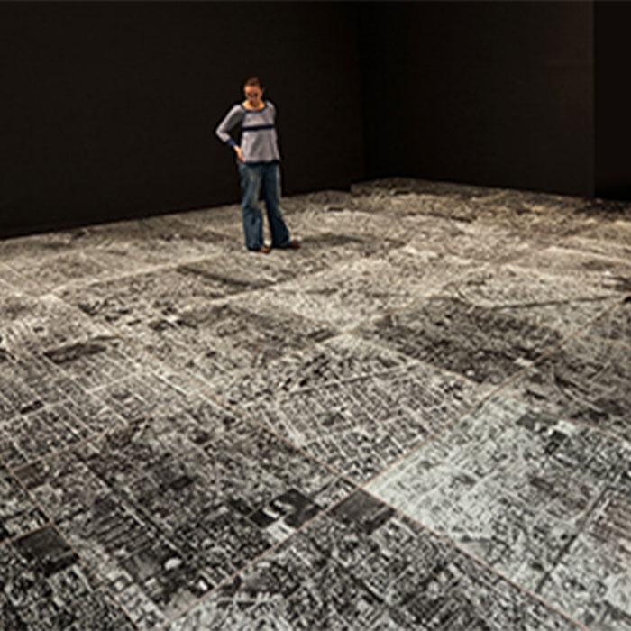 Person standing on Oscar Muñoz's "Ambulatorio (Walking Place/Outpatient Ward)" a floor piece consisted of several tiles of encased broken glass on top of a black and white photograph of Cali Columbia