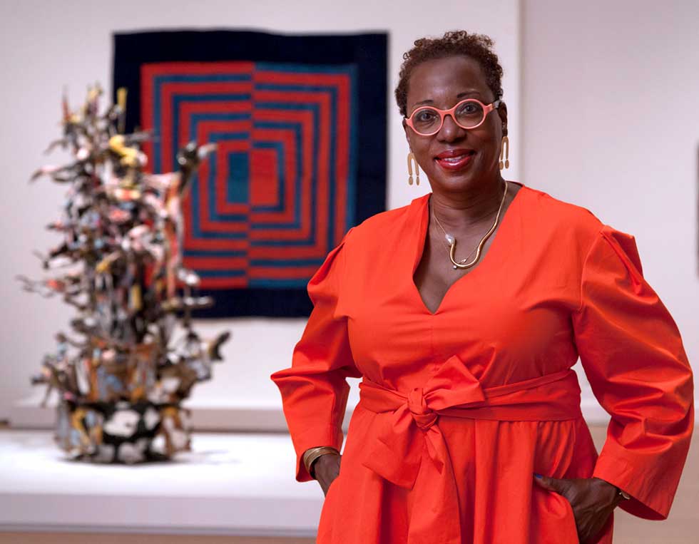 Photo of Valerie Cassel Oliver in a red dress standing in a galley with a sqaure work on the wall and sculpture behind her