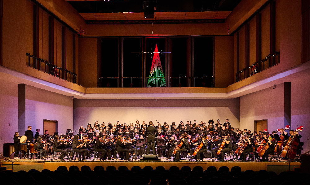 musicians performance on stage in holiday attire, with a lighted tree above them 