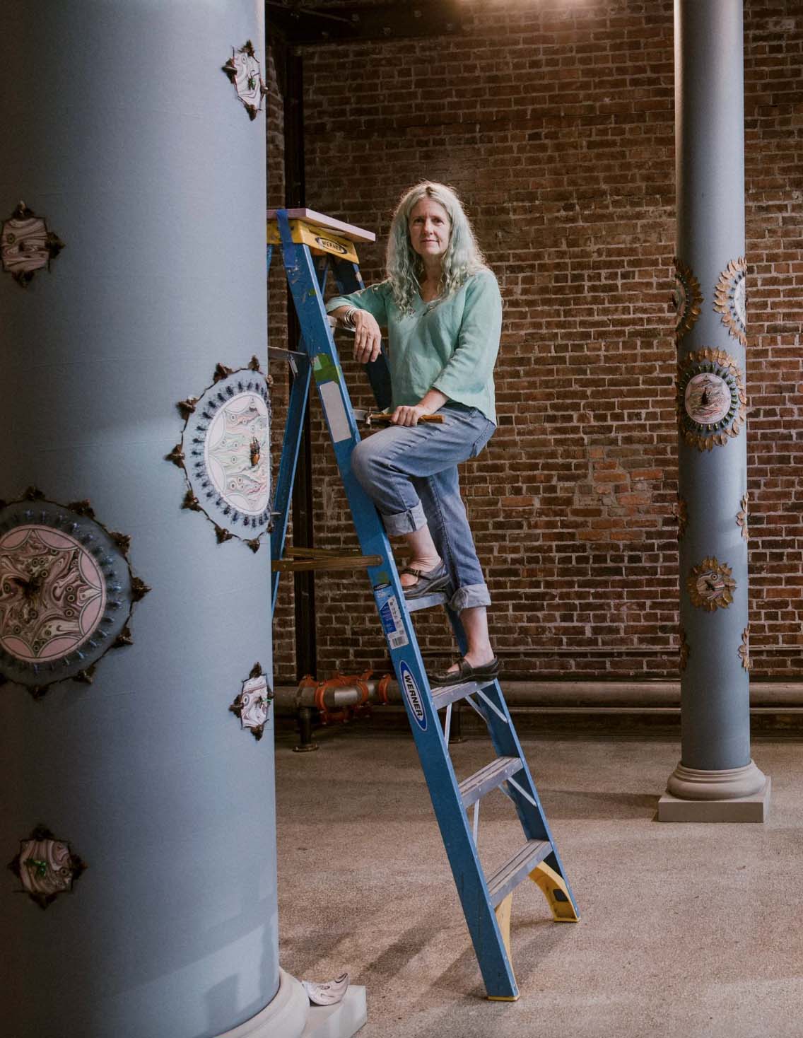 woman on ladder next to columns with artwork
