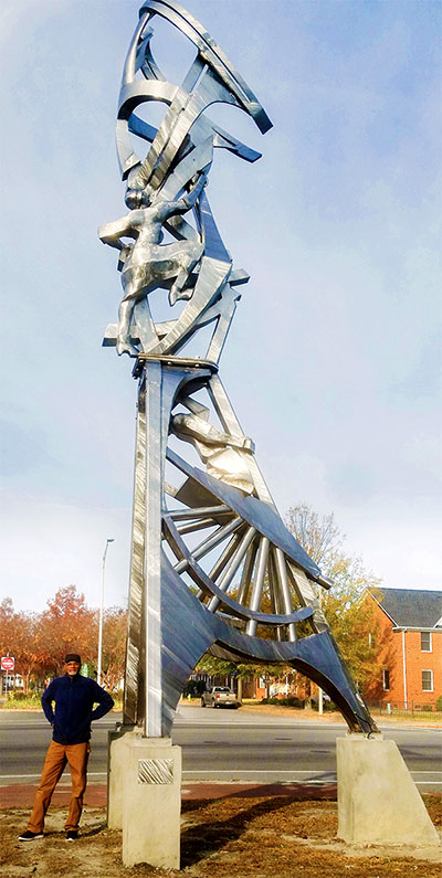 arts professor stands next to his tall, abstract metallic sculpture  