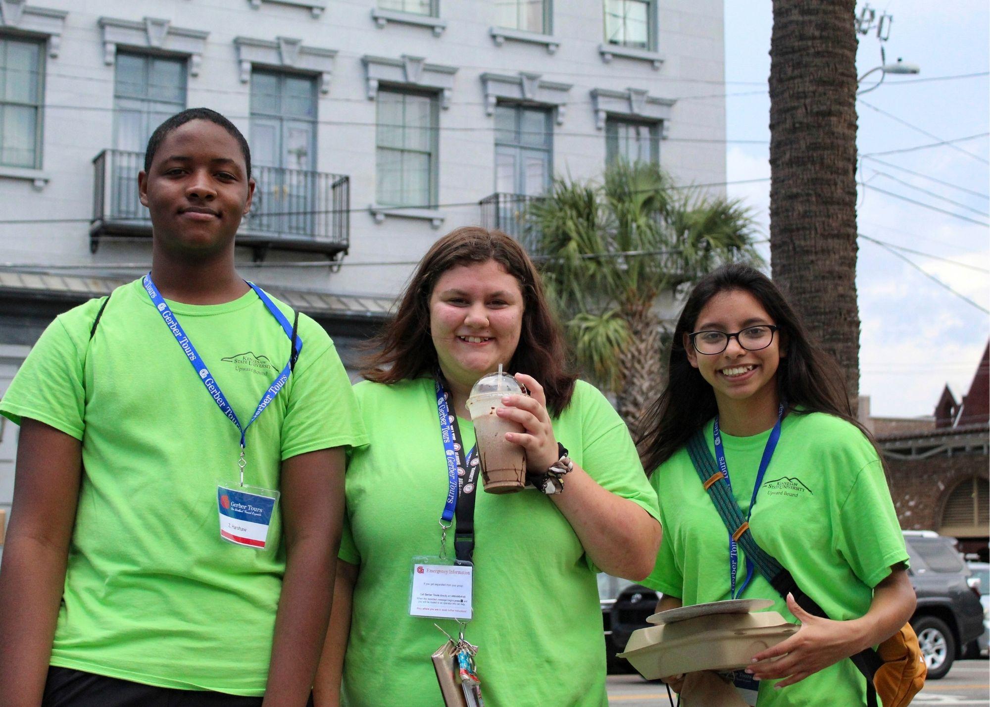 Three students smiling, wearing green shirts standing outside in front of a white building. / KSU TRIO Students have the opportunity to go on an out of state, educational field trip after full participation in the KSU TRiO Summer Academy. The trip is provided at no cost to students.