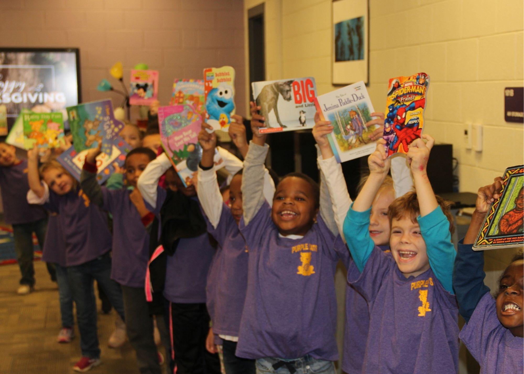 Students dressed in purple shirts smiling and holding books in the air. / Students and staff from the Kennesaw State University and the Georgia Highlands College Paulding Site provided a special Book Drive for Villa Rica students. The groups went to Villa Rica Elementary School with hundreds of free books in order to give students access to a variety of reading material to build their reading skills.