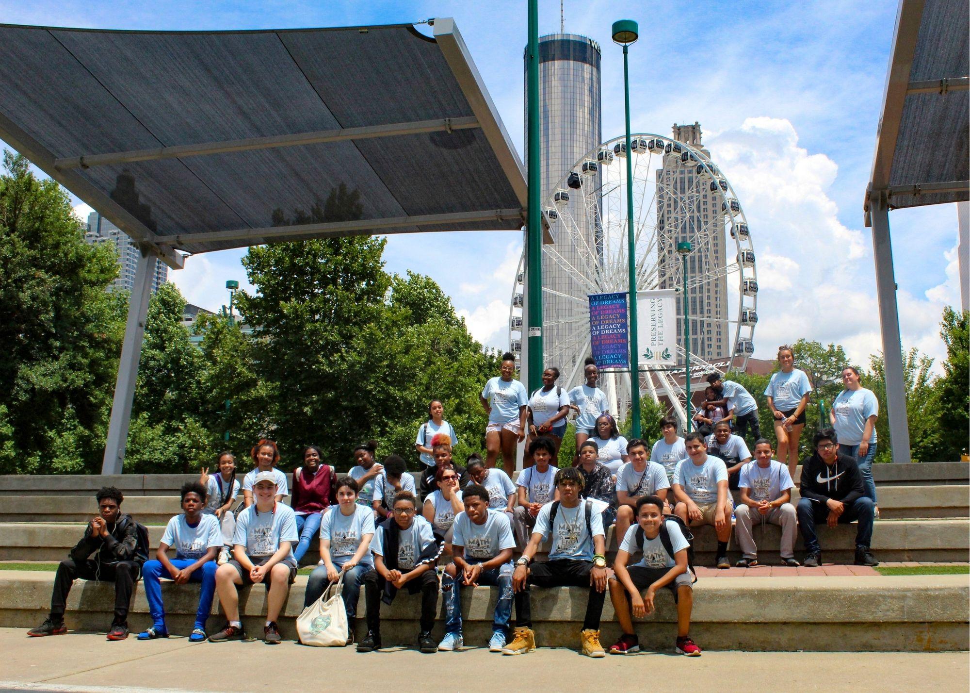 Students wearing blue shirts posing outside in front of a Ferris wheel. / KSU TRiO visits Georgia State University, the Civil Rights Museum, and Centennial Park.