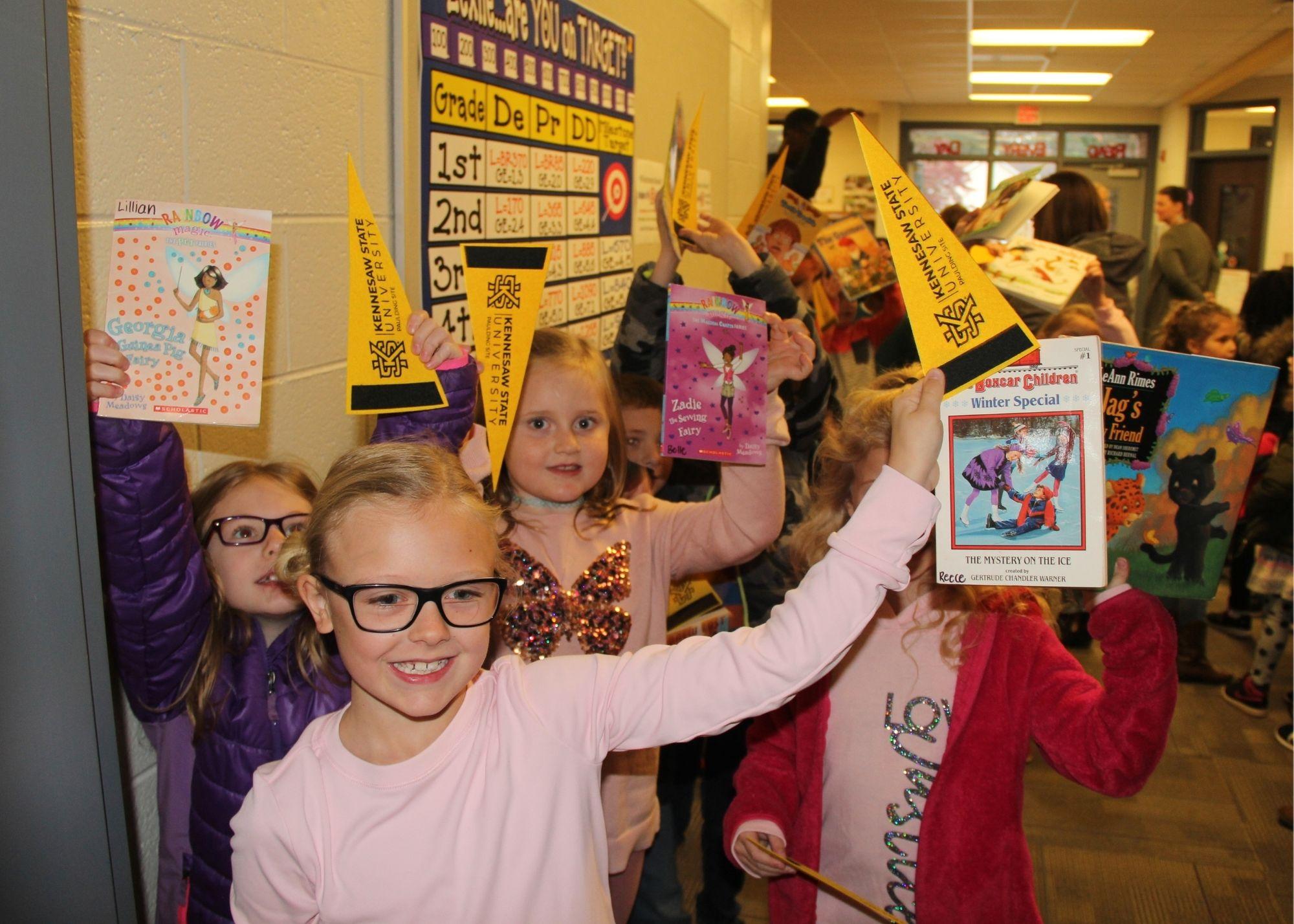 Young students in a hallway holding up books and school flags / Students and staff from the Kennesaw State University and the Georgia Highlands College Paulding Site provided a special Book Drive for Villa Rica students. The groups went to Villa Rica Elementary School with hundreds of free books in order to give students access to a variety of reading material to build their reading skills.