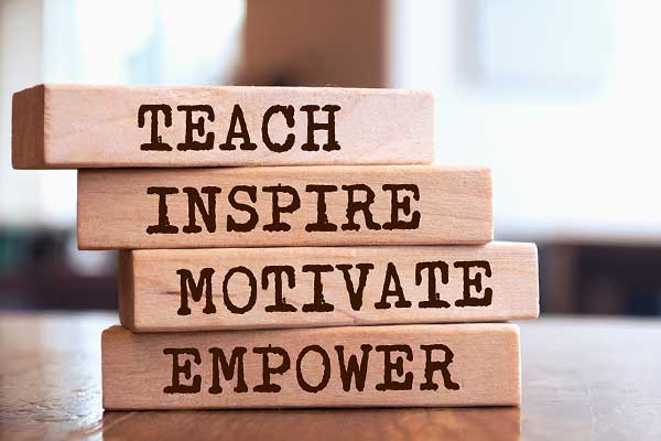 Four wooden blocks are stacked vertically. Each block has a word written in bold, uppercase letters. From top to bottom, the words are "TEACH," "INSPIRE," "MOTIVATE," and "EMPOWER." The background is blurred, drawing attention to the blocks.