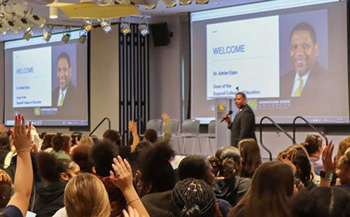 Photo of a large Aspiring Educators of Tomorrow Conference with attendees actively participating, raising their hands in engagement.