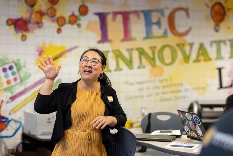 An instructor in a mustard dress and black cardigan gestures while speaking in a colorful "ITEC INNOVATIVE LAB" classroom.