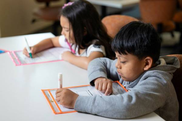 Two young students siting at a table holding markers and doing a writing exercise.