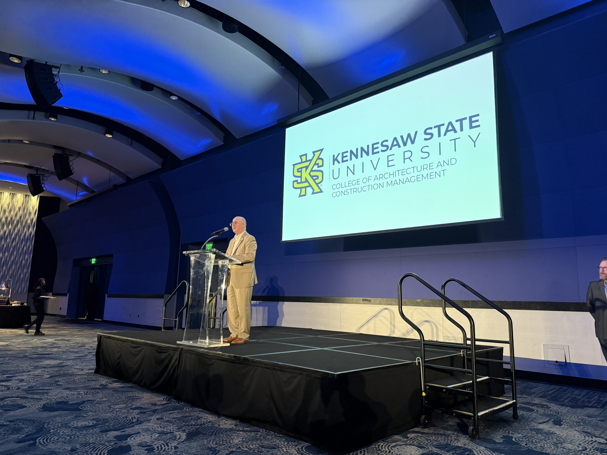 Professor Brian Moore stands on stage in the GA Aquarium's Oceans Ballroom in front of the KSU logo. 